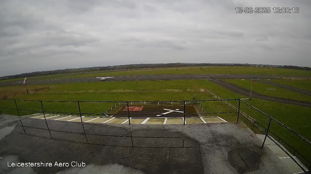 A wide view of a grassy airfield under a cloudy sky. In the foreground, there is a railing from a viewing area, with a few white stripes visible on the ground. A small airplane is seen taxiing on the runway in the background. The runway is dark and paved, bordered by a fence. The scene is tranquil, indicative of an aviation area.