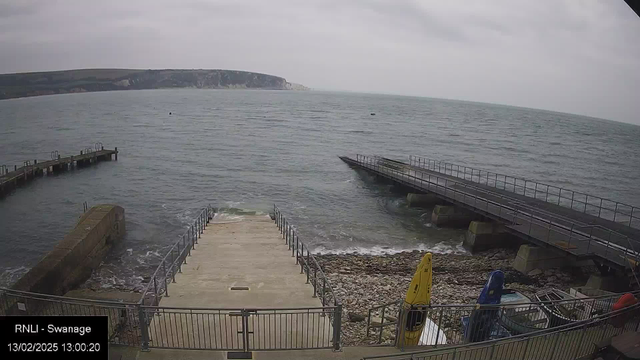 The image shows a coastal scene with a view of the ocean under a gray sky. In the foreground, there is a boat ramp leading down to the water, bordered by a railing. Several boats, including yellow and blue kayaks, are visible on the right side, secured in a storage area. To the left, a stone structure extends into the water. The ocean appears calm, with small waves lapping at the rocky shoreline. In the distance, cliffs are seen along the coastline. The date and time are displayed in the bottom left corner, indicating it is February 13, 2025, at 13:00:20.