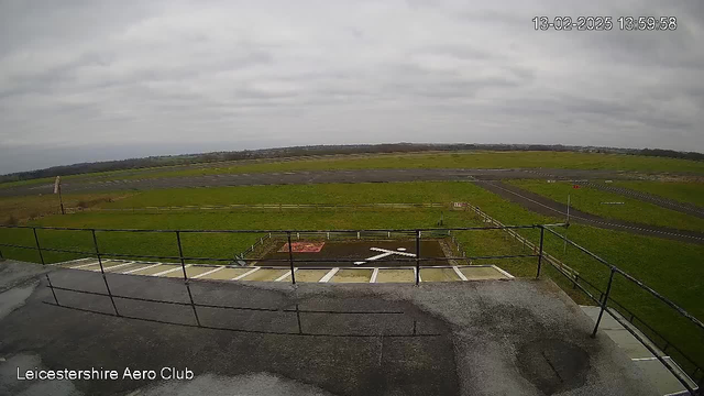 A view from the top of a building at an aero club, overlooking a grassy airfield. The sky is overcast with clouds. In the foreground, there is a railing along a flat rooftop, leading down to a ramp. A large white cross is marked on the ground in the airfield below, which is surrounded by an open grassy area. A small building and some fences can be seen in the distance, along with a tarred runway.