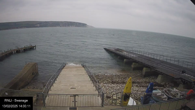 A cloudy day over a coastal scene, featuring a calm sea. In the foreground, a wide concrete ramp descends toward the water, bordered by a fence. To the right, there is a dock with a ramp extending into the water. Alongside the concrete area, several kayaks are positioned on the shore with colors including yellow, blue, and red. The shoreline is rocky, with pebbles visible in the water's edge. In the background, a green hillside rises sharply along the coast, leading to cliffs under the overcast sky.