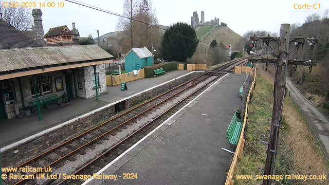 An empty railway station platform with stone walls and a green roofed building. Several green benches line the platform. In the background, there are hills with castle ruins visible at the top. A wooden post with electrical wires is positioned on the right side. The sky is cloudy, creating a gray atmosphere. A sign on the platform reads "WAY OUT". There are also several trees and a fenced area on the left side. The tracks run alongside the platform and are lined with gravel.