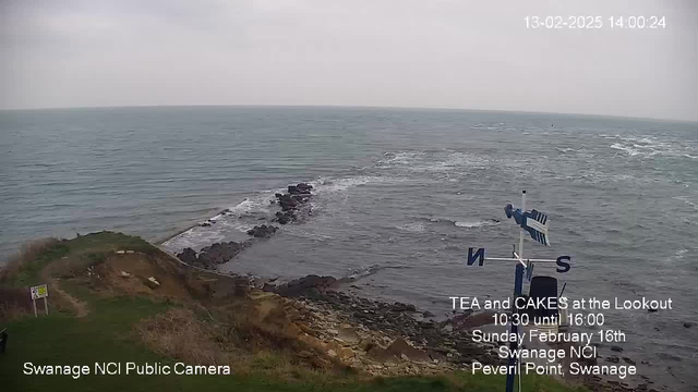 A coastal scene captured in a webcam image features a view of the sea under a cloudy sky. In the foreground, there is greenery and rocky formations along the coast. A tall wind vane with a blue and white striped design is visible, indicating wind direction. The water appears to have small waves, and there are scattered rocks in the ocean. Text on the image promotes a tea and cake event at a lookout point, giving details about the time and location. The atmosphere is calm and slightly overcast.