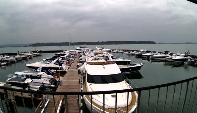 A view of a marina filled with numerous boats. The scene is captured under a cloudy sky, with a calm water surface reflecting the light. Several boats are moored closely together, with a prominent white yacht in the foreground. Wooden docks extend into the water, and distant land is visible along the horizon. The atmosphere is serene, with no visible human activity.