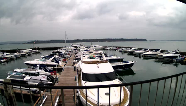 A view of a marina filled with numerous boats docked in still water. The boats are varied in size and design, with several larger yachts prominent in the foreground. The scene is set against a cloudy sky, and a distant landmass is visible across the water. A wooden dock is visible in the foreground, suggesting easy access to the boats. The overall atmosphere is calm and serene.