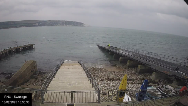 A view of the coastline with a cloudy sky. In the foreground, there is a concrete ramp leading down to stony beach areas and the water's edge. To the left, a wooden pier extends over the water, and in the distance, the coastline features cliffs. On the right side, a person in a bright yellow jacket appears on a metal ramp that leads out to the water. Several kayaks in yellow and blue are positioned nearby on the shore.