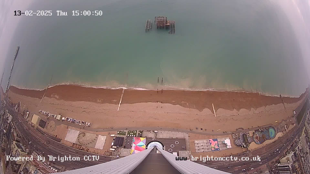 A bird's-eye view of a beach and coastline. The sandy beach stretches across the lower part of the image, bordered by a light brown strip of sand near the water. The turquoise sea meets the shore, and some wooden structures are visible in the water, possibly remnants of a pier. Above the beach, various structures and amenities can be seen along the promenade, including colorful tents and a round attraction. The sky appears overcast. The image is timestamped with the date and time at the top and the source of the webcam at the bottom.