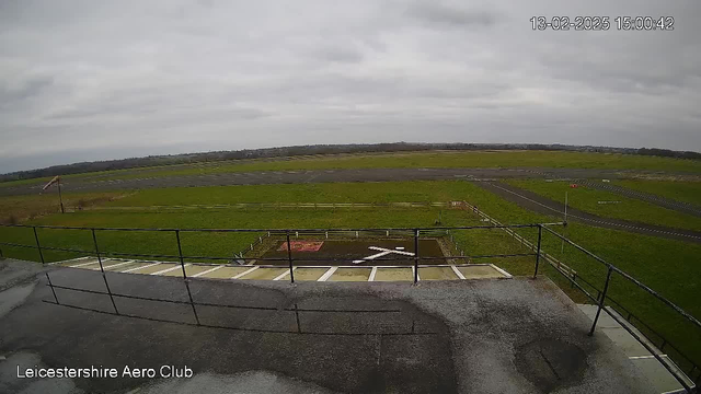 A view from a high vantage point overlooking an airfield. The sky is mostly cloudy and gray. In the foreground, there is a railing and a small platform. Below, a grassy area is visible with a red square and a white cross marking on the ground. The airfield extends into the distance, with a paved runway and some distant fields.