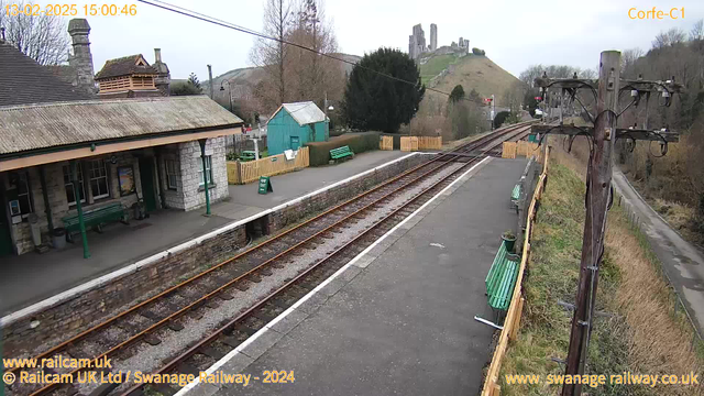 A view of Corfe Castle railway station with a single track running through the center. The station building, made of stone, features a sloping roof and green accents. On the platform, there are several green benches. To the left, there's a small wooden shed painted turquoise and a sign indicating "Way Out," while a wooden fence surrounds an area with more seating. In the background, the ruins of Corfe Castle stand on a hill, partially obscured by trees. The sky is overcast, giving a muted light to the scene.