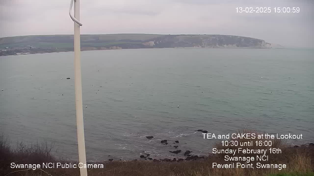 A view from a webcam showing a coastal scene at Peveril Point, Swanage. The image features grey skies and a calm sea with slight waves. In the foreground, there are some rocks along the shoreline, and small boats can be seen in the water. The coastline is visible in the distance, with grassy hills leading to cliffs. Text in the lower right corner announces a tea and cake event at the Lookout on February 16th, 2025.