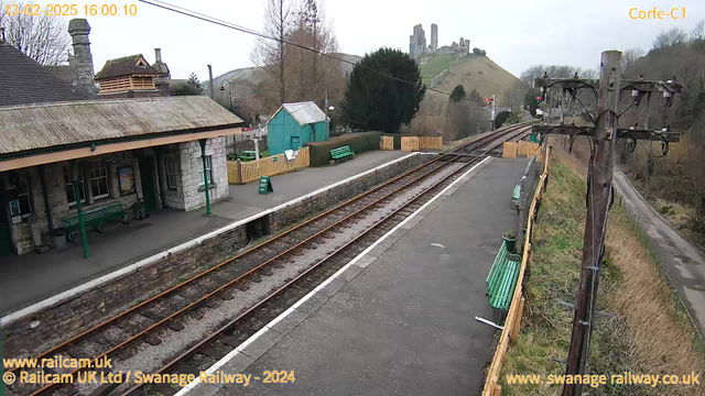 A view of Corfe Castle railway station on a cloudy day. The image shows a stone building with a peaked roof and a covered porch area. Green benches are placed along the platform, and there is a small turquoise shed in the background. The tracks are visible, leading away from the platform, bordered by a low stone wall. In the distance, Corfe Castle can be seen atop a hill, surrounded by trees. A wooden fence encloses a grassy area adjacent to the platform. Utility poles with wires are present on the right side of the image.