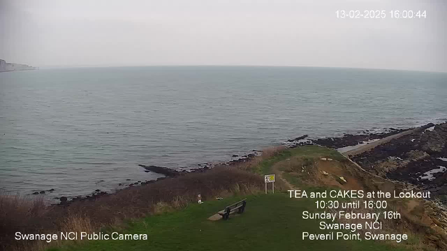 A coastal scene showing the ocean and rocky shoreline. The water is calm and a light gray sky is above. In the foreground, there is a grassy area with a wooden bench facing the sea. A sign nearby announces a tea and cakes event, mentioning the time and date. The shoreline includes dark rocks and scattered vegetation.