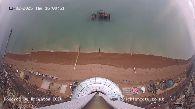 A high-angle view of a sandy beach with a calm blue-green sea. The beach has a few wooden piers extending into the water, and several dark, abandoned pier structures visible offshore. To the right, there are beachside attractions, including a pool and colorful patterns on pavement. The scene is under an overcast sky, with a portion of a building's circular glass dome at the bottom of the image. The image is timestamped with the date and time at the top.