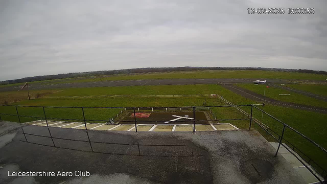 A view from a webcam at Leicestershire Aero Club shows a cloudy sky over an airport. A small aircraft is taxies on a runway to the right, while empty patches of grass and a dirt area with a white cross marking are visible in the foreground. The landscape is generally flat, with distant trees lining the perimeter of the airfield. There are signs and a boundary fence along the edge of the runway.