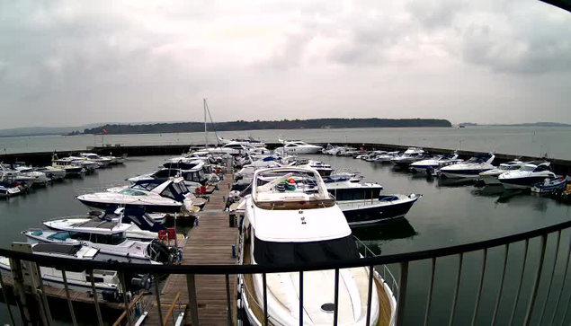 A view of a marina filled with numerous boats docked in a calm body of water. The scene is overcast with gray clouds, and a wooden pier extends into the water. Boats are clustered together, some with colorful covers or accessories visible on their decks. In the distance, a shoreline can be seen, and the water reflects the cloudy sky.