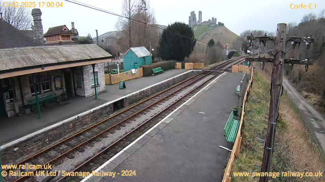 A view of a railway station platform at Corfe Castle. The platform features a stone building with a sloped roof, green benches, and a noticeboard. In the background, there is a train track leading to the right, with two tracks visible. To the left, a green shed and wooden fencing surround the area. A distant hill can be seen with the ruins of a castle on top. The sky is overcast, and the scene appears calm and quiet.