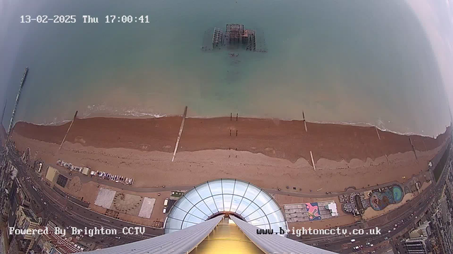 A bird's-eye view of a beach scene. The image shows a sandy beach with waves gently lapping at the shore. In the foreground, there is a circular glass structure, possibly a viewing platform, with tall supports extending downwards. Below the water, remnants of an old pier can be seen. Along the beach, there are hints of amusement attractions, likely rides, and colorful structures. The sky above is cloudy, and the time and date are displayed at the top of the image.