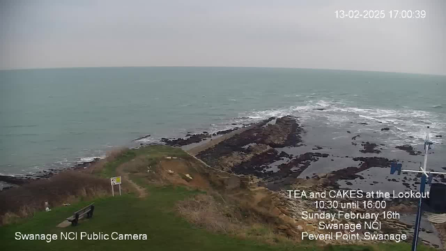 A coastal scene showing the sea with gentle waves and a rocky shoreline. In the foreground, there is a grassy area with a bench and a sign. The sky is overcast, creating a muted atmosphere. The time and date are displayed in the upper right corner, along with information about a community event at the Lookout, indicating it takes place on February 16th, between 10:30 AM and 4:00 PM, at Peveril Point, Swanage.