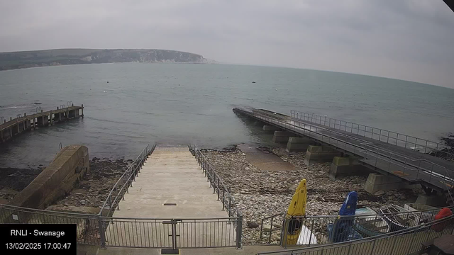 A coastal scene shows a view of the sea under a cloudy sky. In the foreground, there are steps leading down to the water, flanked by a metal railing. To the left, there is a wooden pier extending into the water. The shoreline is rocky, with a few scattered boulders and a small sandy area visible. Several kayaks in different colors are stored near the bottom right of the image, while the sea appears calm with a few distant boats. The cliffs can be seen in the background on the opposite side of the water. The time and date at the bottom indicate it is February 13, 2025, at 17:00:47.