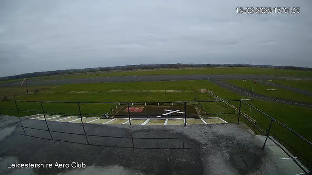 A wide view of an airfield taken from an elevated position. The foreground shows a railing with a flat surface below. In the middle ground, there is a grassy field with a few light poles and a runway marked with white lines. In the background, the sky is overcast with gray clouds, and the landscape fades into distant greenery. The date and time display is in the upper right corner of the image.