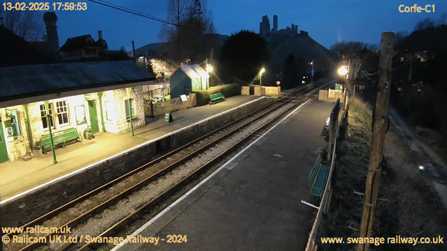 A railway station is visible at dusk, illuminated by street lights. The platform features green benches and a green sign saying "Way Out." A stone building with several windows is present, and a light illuminates the area. In the background, hilltop ruins are silhouetted against the darkening sky, with the railway tracks running parallel to the platform. The scene conveys a quiet, tranquil atmosphere.