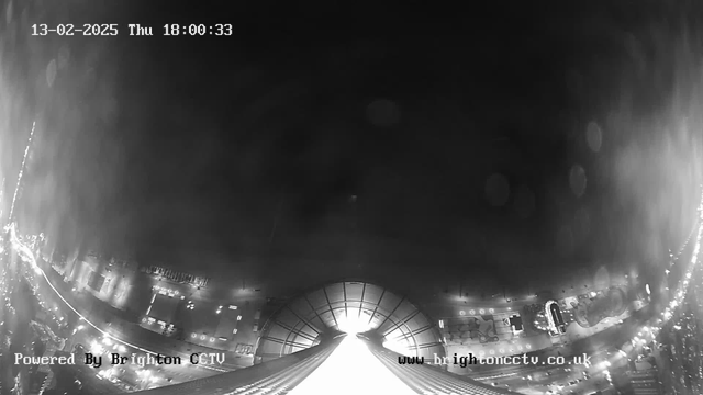 A high-angle view captured by a webcam, featuring an urban scene at dusk. The image is predominantly dark with faint light streaks suggesting traffic below. The horizon stretches across the lower part of the image, revealing outlines of buildings and streets lit by artificial lights. The top of the frame is lighter, indicating a bright area, potentially the sky, transitioning to dark above. The date and time, "13-02-2025 Thu 18:00:33," are displayed at the top, with "Powered By Brighton CCTV" in the lower left corner and a web address at the bottom right.