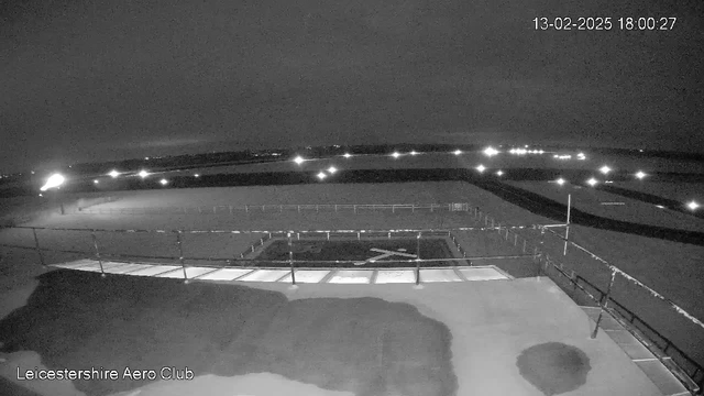 A dark nighttime scene captured from a webcam at Leicester Aero Club. In the foreground, a partially illuminated flat area can be seen, along with a railing and what appears to be a flat surface with faint markings. The background consists of a dark sky with scattered bright spots indicating distant lights. The overall atmosphere is dim, with minimal visibility and a slight overcast. The image is timestamped: February 13, 2025, at 18:00:27.