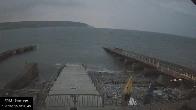 A view of a rocky shoreline at dusk, with calm gray water extending to the horizon. In the foreground, a concrete ramp leads down to the water, bordered by metal railings. To the right, a wooden pier extends out into the water. On the right side of the image, several boats are visible, including a yellow kayak. The background features cliffs and a cloudy sky.