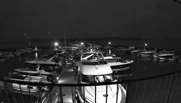 A nighttime scene of a marina filled with various boats docked along a wooden pier. Some boats are illuminated by nearby lights, creating reflections on the dark water. The area is mostly quiet, with a few dim lights in the background. The sky is dark, contributing to the overall ambiance of a peaceful harbor.