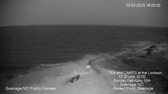 A coastal scene is visible under low light, showing a vast expanse of dark water and waves gently lapping onto a rocky shoreline. In the foreground, there are some rocks and a small structure that appears to be a bench. The sky above is overcast, contributing to the muted lighting. Text on the image indicates the date and time, along with information about a local event at a lookout point, which is also noted at the bottom left.