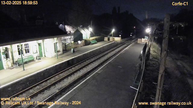 A dimly lit railway platform at night, featuring a stone building with green doors and multiple windows on the left side. A green bench is positioned near the building. There are wooden barriers along the platform, with another green bench visible further along. Two illuminated lamps provide light on the platform. The railway tracks run parallel to the platform, with gravel visible between the tracks. The scene conveys a quiet, empty atmosphere typical of a railway station at night.