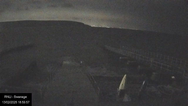 A dimly lit scene depicting a coastal area at night. In the foreground, there is an incomplete structure or dock with some scattered objects, including what appears to be a white sail and some wooden beams. To the right, there is a railing leading away toward the horizon. In the background, faint lights from a distant town or city can be seen against a cloudy sky. The overall atmosphere is quiet and somewhat mysterious due to the low light levels.