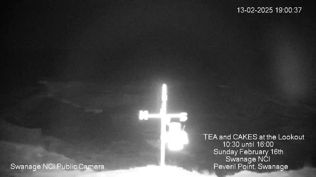 A black and white webcam image showing a cross structure with directional markers indicating North, South, East, and West. The background is a dark, cloudy sky with hints of a coastline visible near the bottom. There is text at the bottom left stating "Swanage NCI Public Camera" and additional details about an event titled "TEA and CAKES at the Lookout" taking place on Sunday, February 16th, from 10:30 to 16:00 at Swanage NCI, Peveril Point, Swanage. The date and time in the top right corner indicate "13-02-2025 19:00:37."