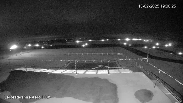 A night scene from a webcam at Leicestershire Aero Club showing a dark landscape. The image features a wide, empty airfield with faint lights in the distance. A portion of the foreground includes a railing or fence, and there are patches of light reflecting off a surface, suggesting a structure or pathway. The date and time displayed at the top right indicates it is February 13, 2025, at 19:00:25.