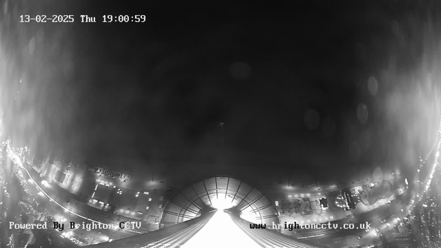 A black and white image from a webcam showing an urban landscape at night. The scene is mostly dark, with faint outlines of buildings and streets. There are light spots that suggest streetlights or vehicle headlights. The bottom part of the image shows a perspective looking upward, leading to a bright area at the center, which might indicate a bright opening or structure. A timestamp in the upper left corner reads "13-02-2025 Thu 19:00:59," and the bottom left corner has the text "Powered By Brighton CCTV." The overall mood is quiet and somewhat empty, characteristic of a nighttime city view.