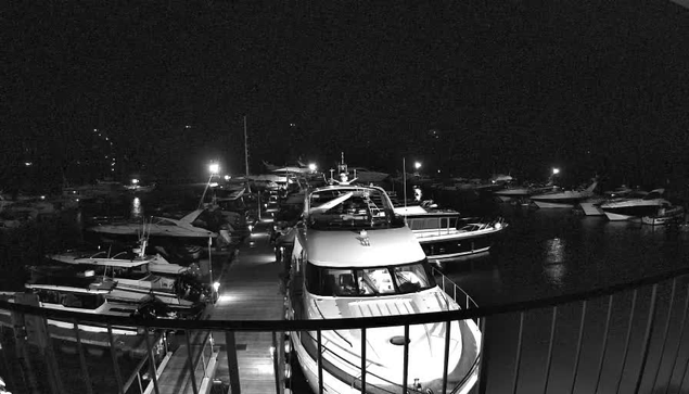 A dark scene of a marina at night, filled with various boats and yachts moored along a wooden dock. Several boats are illuminated by artificial lights, casting reflections on the water's surface. The overall atmosphere is quiet, with some light sources in the background indicating activity in the area.