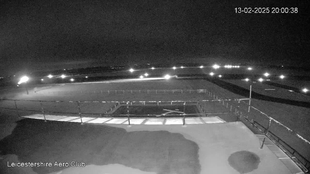 A nighttime view from a webcam at Leicestershire Aero Club. The image is mostly dark, with some illuminated areas on the ground. There is a runway with lights lining it, and a cross made of lights is visible on the tarmac. The surrounding area appears to be open land with minimal structures. The date and time are displayed in the corner.