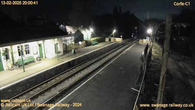 A dimly lit railway station at night, featuring a stone platform with green benches. The platform is empty, and a pair of railway tracks runs through the scene. There are lampposts providing some light, with the illumination casting soft shadows. In the background, a building with multiple windows is partially visible, along with some greenery. A sign reading "WAY OUT" is placed on the platform. The setting conveys a quiet, peaceful atmosphere typical of a rural station at night.