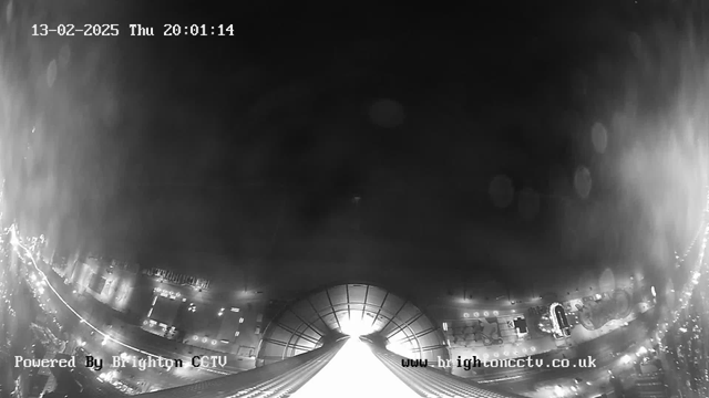 A black and white image from a webcam shows a wide view of a city at night. The scene is dark with some lights visible along the bottom, likely from buildings and streetlights. A bright light source appears in the center, possibly indicating an illuminated area. The date and time are displayed in the top left corner as "13-02-2025 Thu 20:01:14," while the bottom part of the image includes the text "Powered By Brighton CCTV" and a website URL. Water droplets or lens artifacts are present, adding to the hazy appearance of the image.