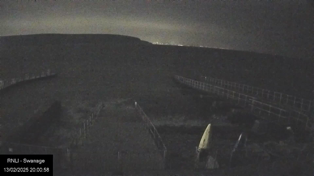 A dark, nighttime scene showing a coastal area. In the foreground, there are outlines of a few objects, possibly boats or kayaks, with one yellow object prominently displayed. A wooden walkway or jetty is partially visible, leading off into the darkness. The background features a distant, soft glow of light, indicating a town or city on the horizon, surrounded by a dark hill silhouette. The overall atmosphere is quiet and tranquil, indicative of a seaside location at night.