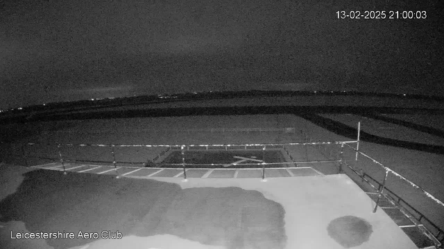 A dark scene captured by a webcam at Leicester Aerospace Club, showing a flat surface with faint markings, possibly a tarmac area. In the background, there are scattered lights indicating the presence of distant buildings or structures. The sky is overcast with minimal illumination, suggesting it is nighttime. The date and time stamp in the corner reads "13-02-2025 21:00:03."