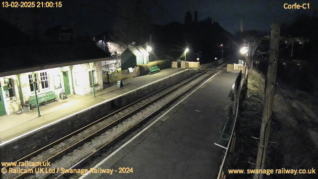 A dark scene at a railway station. The platform is mostly empty, with a few green benches and a sign that reads "WAY OUT." There is a stone building on the left with large windows and an awning. Soft light illuminates the area, creating a calm atmosphere. The tracks are visible, leading into the darkness, with wooden fencing along the edge of the platform. In the background, there are silhouettes of trees and possibly buildings. The date and time displayed read 13-02-2025, 21:00:15.