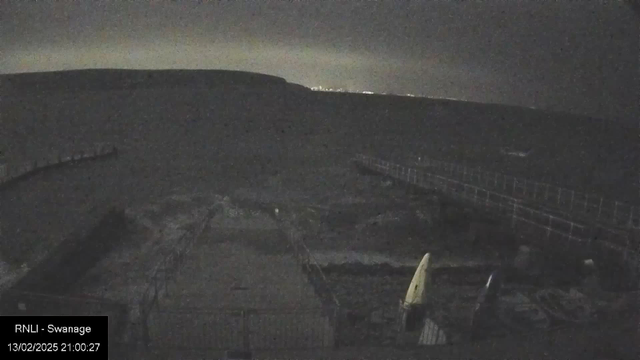 A dark, nighttime scene of a waterfront area. The foreground shows a rugged surface with a few objects, including a yellow boat and some other equipment. There is a visible railing along a path leading to the water, which is partially illuminated by distant lights on the horizon. The sky is overcast, contributing to the overall dim atmosphere.