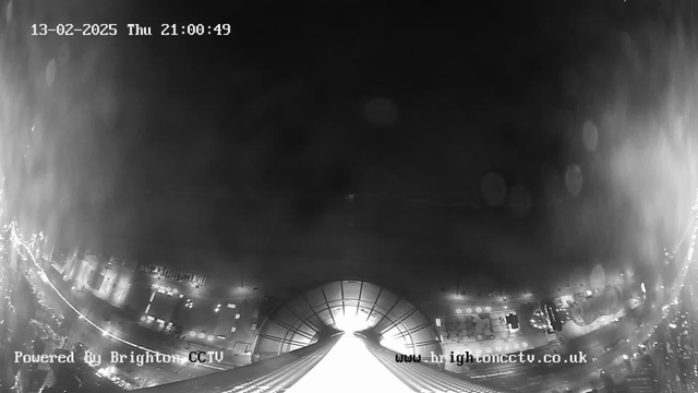 A black and white webcam image shows a view from a high vantage point, looking down towards a city street below. The foreground features a bright, illuminated area that appears to be a structure or roof edge leading into darkness. In the distance, faint outlines of buildings and streetlights are visible, suggesting a cityscape. The image contains a timestamp at the top left corner indicating the date and time. There are blurred spots likely caused by raindrops or moisture on the lens. The bottom section includes text indicating it is powered by Brighton CCTV.