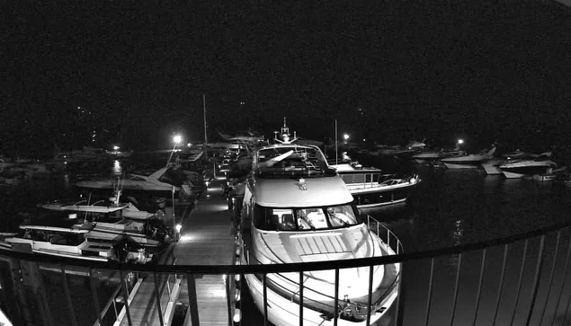 A nighttime view of a marina filled with various boats docked along a wooden pier. The scene is illuminated by several bright lights reflecting on the water, creating a shimmering effect. The boats vary in size and type, with some showing visible details like windows and railings. The overall atmosphere is calm and still, with dark waters surrounding the vessels.