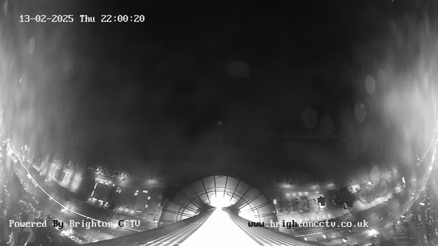 A black and white image from a webcam, showing a nighttime cityscape. The view is slightly angled downwards, revealing illuminated streets and buildings below. The lower part of the image features a section of a curved rooftop or structure leading to a bright area in the center, likely illuminated by streetlights. The date and time are displayed at the top, indicating February 13, 2025, at 22:00:20. A watermark at the bottom left indicates the image is powered by Brighton CCTV.