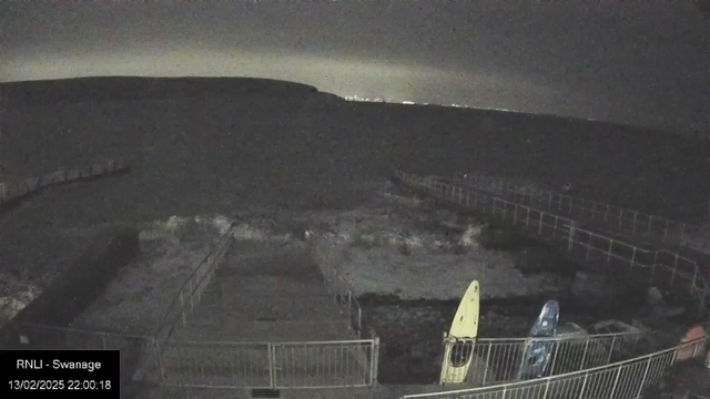 A nighttime scene showing a waterfront area with a visible pier extending into the dark water. There are two kayaks on the shore, one yellow and one blue, positioned next to a railing. The background features indistinct hills and a faint glow of distant lights on the horizon. The overall atmosphere is dark with a few illuminated areas.