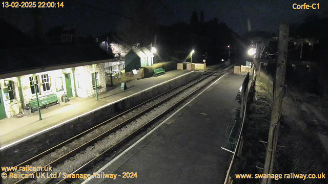 A dimly lit train station platform at night, featuring a stone building with several windows. There are wooden benches on the platform and a sign that reads "WAY OUT." The tracks are visible, leading away from the platform, and there are fenced areas on the right. The scene is illuminated by a few artificial lights, with trees and a building silhouette in the background.