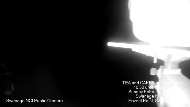 A dark image with a bright light source on the upper right, casting a strong glare. The left side features the outline of an object, possibly a railing or a pole. At the bottom, text includes "Swanage NCI Public Camera" and details about an event: "TEA and CAKES at the lookout 10:30 until 1:00, Sunday February 19, Swanage NCI, Peveril Point, Swanage." The overall scene feels dim, with the focus primarily on the bright light and the text below.