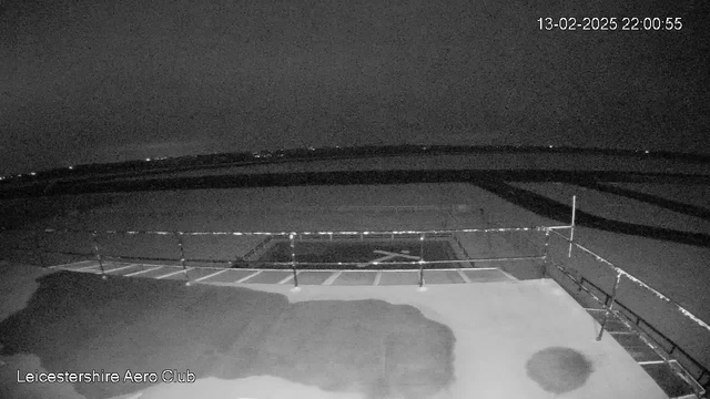 A dark nighttime scene from a webcam overlooking an airfield. In the foreground, there's a section of ground with uneven surfaces and shadows. A barrier is visible, indicating the edge of a platform or building. In the distance, faint lights are scattered on the horizon, possibly indicating nearby structures or roads. Overall, the image has low visibility with a monochromatic color scheme.