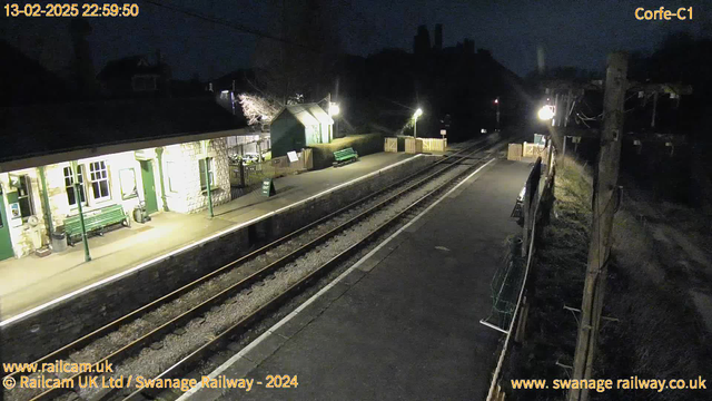 A dimly lit train station at night, showing two railway tracks converging towards the horizon. On the left side, there is a stone building with large windows and a bench outside. The building is illuminated, revealing its structure and some details. Nearby, there are green benches and a sign indicating "WAY OUT." To the right, a wooden fence separates the platform from a grassy area. Utility poles with wires are visible in the background, and the atmosphere is quiet and still.